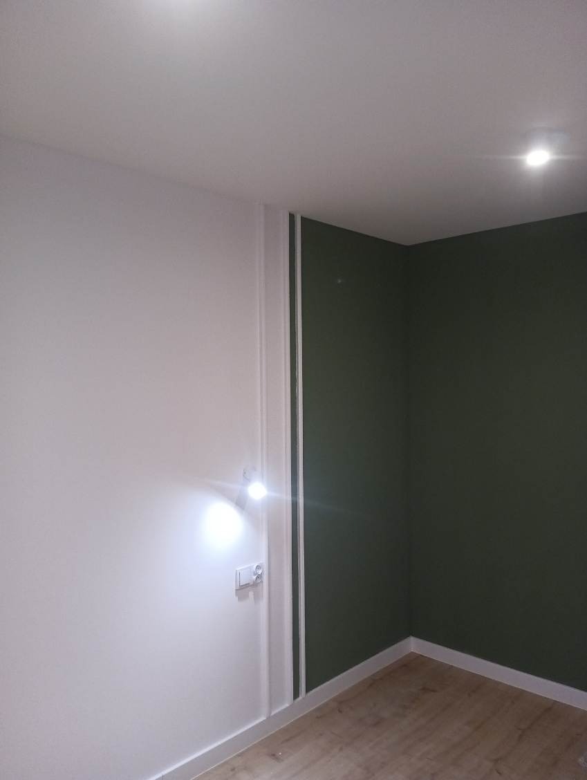 Pomalowane na biało i oliwkowo ściany w rogu pokoju, z widoczną listwą przypodłogową i włącznikiem światła, oświetlone punktowym światłem.