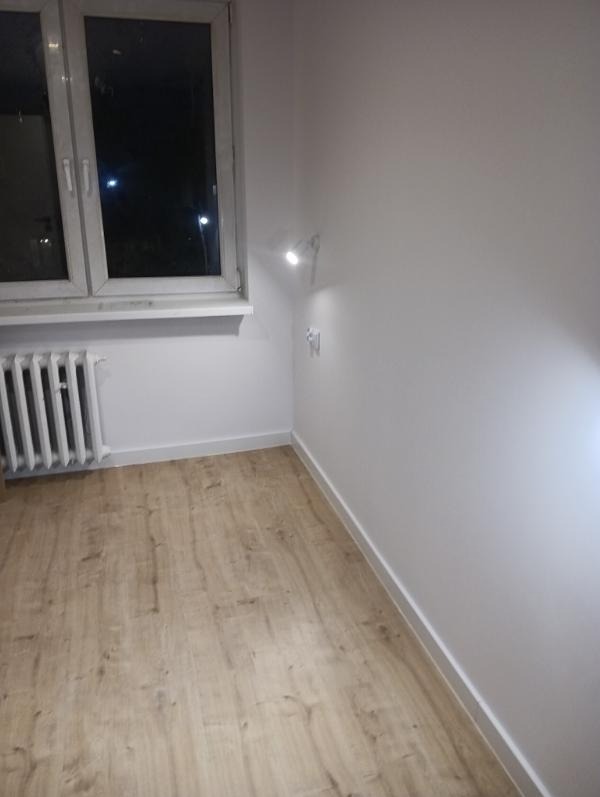 Jasne panele podłogowe w pokoju po remoncie, białe ściany, okno z widokiem na noc, grzejnik i minimalistyczna lampka ścienna. Widok na wykończone wnętrze.