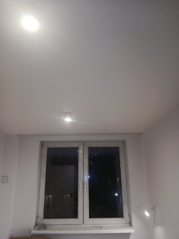 Biały sufit po gładziowaniu i malowaniu, widoczne okno z parapetem. Całość oświetlona od góry i dołu. Minimalistyczne wykończenie wnętrza.