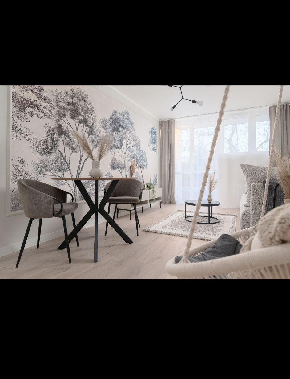 Jasne wnętrze z fototapetą w motywy leśne, okrągłym stołem z krzesłami i wiszącym fotelem. Parkiet w odcieniach beżu. Dekoracyjne dodatki z suszonych traw.