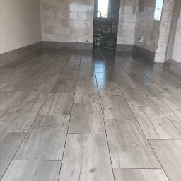 TEMIDA HOME SPÓŁKA Z OGRANICZONĄ ODPOWIEDZIALNOŚCIĄ - Ułożone podłużne płytki podłogowe imitujące drewno w jasnoszarym odcieniu, przy ścianach widoczne płytki ścienne, w głębi pomieszczenia otwarte przejście do innego pomieszczenia z widocznym sprzętem.