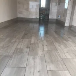 Ułożone podłużne płytki podłogowe imitujące drewno w jasnoszarym odcieniu, przy ścianach widoczne płytki ścienne, w głębi pomieszczenia otwarte przejście do innego pomieszczenia z widocznym sprzętem.