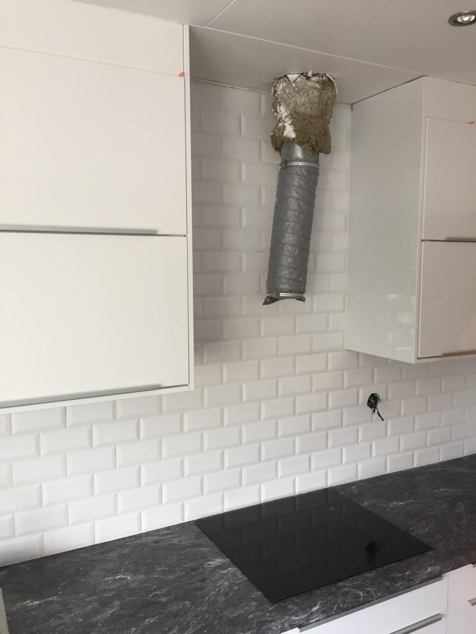 Niedokończony montaż okapu kuchennego z widocznym podłączeniem do wentylacji, białe szafki wiszące i czarny blat z płytą indukcyjną oraz białe płytki typu metro na ścianie.