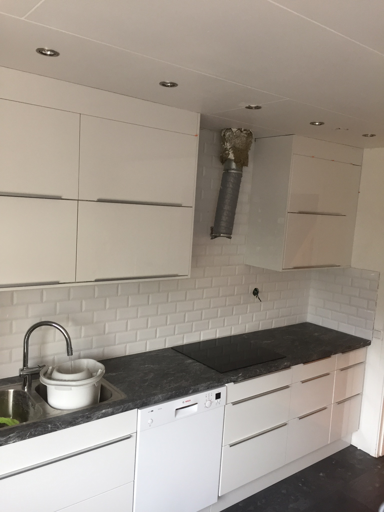 Nowoczesna kuchnia z białymi szafkami, ciemnym blatem i płytkami typu metro na ścianie, widoczna rura wentylacyjna w trakcie montażu i wiadro w zlewie.