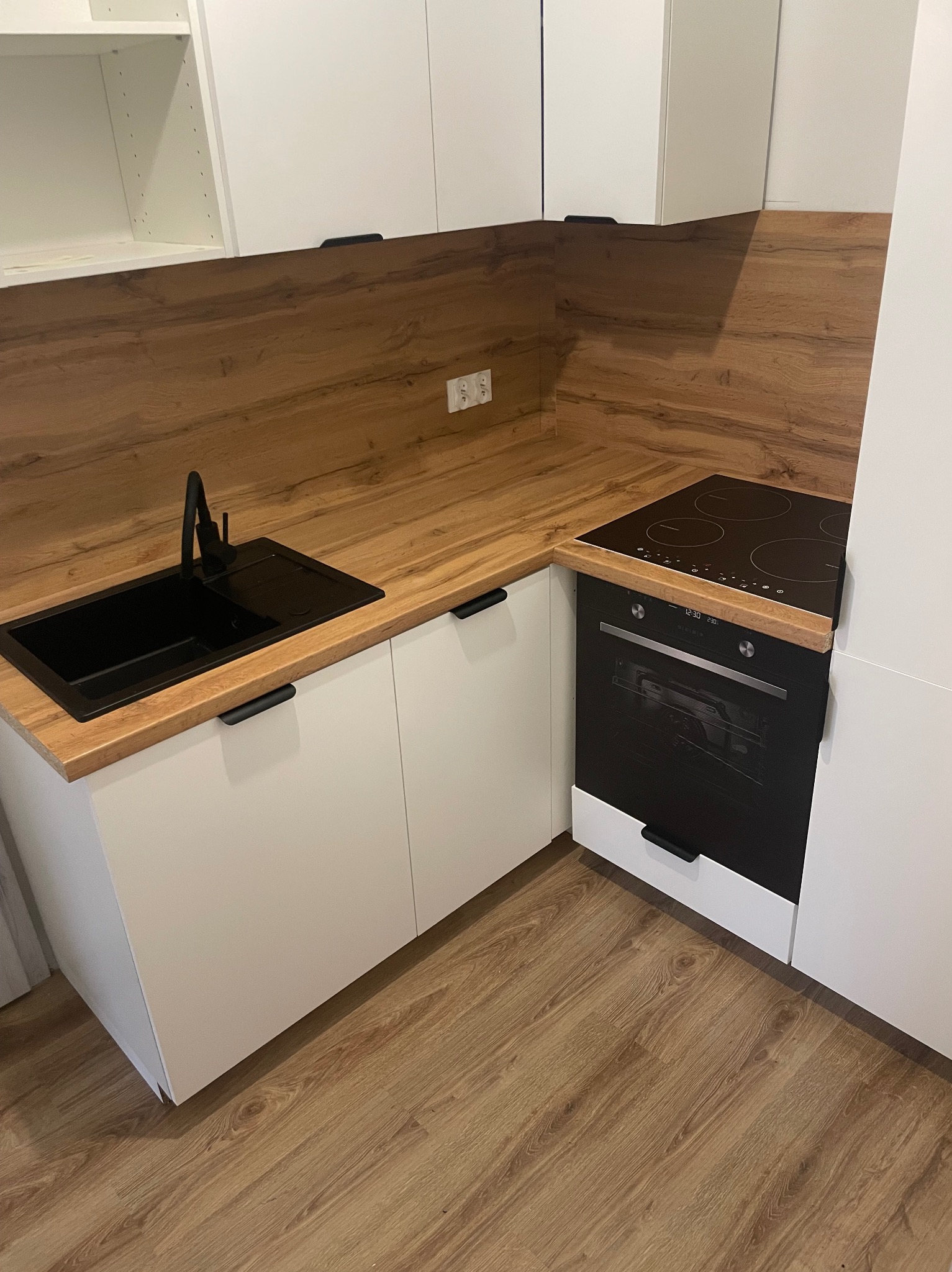 Nowoczesna kuchnia z białymi szafkami, blatem w kolorze drewna, czarnym zlewozmywakiem i płytą indukcyjną z piekarnikiem w zabudowie.