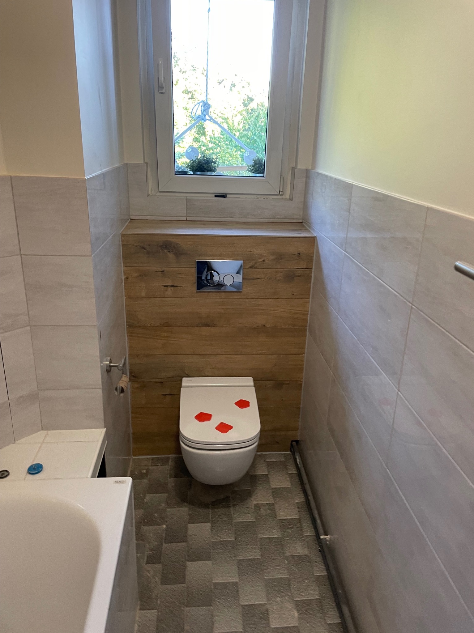 Wykończona łazienka z podwieszaną toaletą, drewnianą zabudową za toaletą, szarymi płytkami podłogowymi i jasnymi płytkami ściennymi, okno z widokiem na zieleń, czerwone płatki na desce toaletowej.