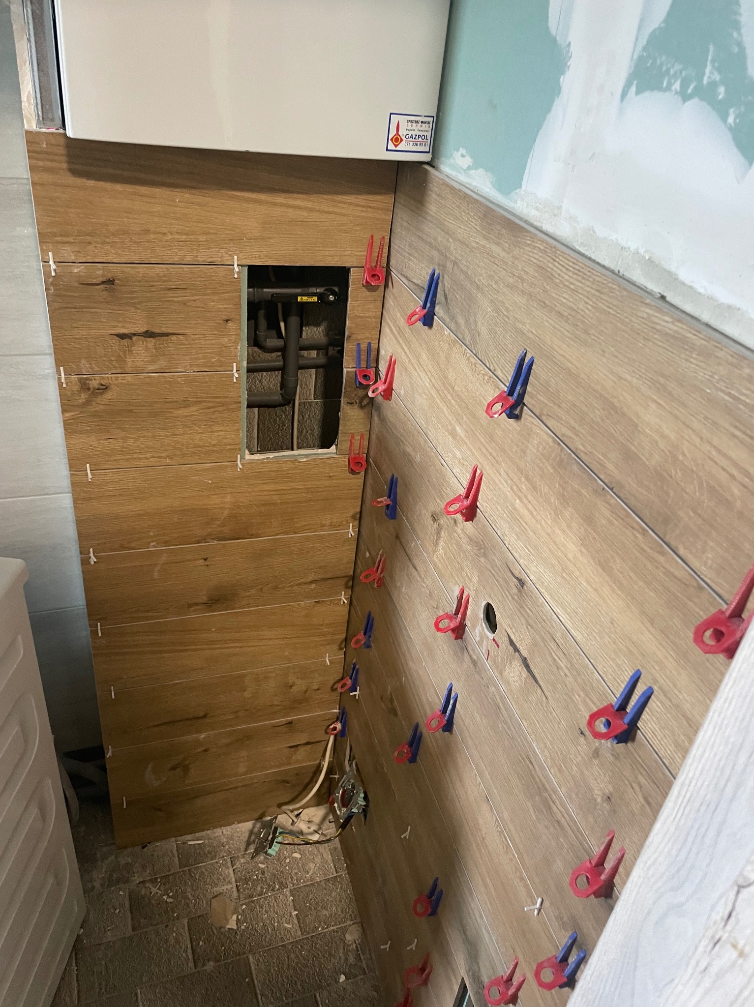 Ściana w trakcie układania płytek imitujących drewno, z widocznymi klipsami poziomującymi w kolorach czerwonym i niebieskim, otwór na rury instalacyjne i fragment kotła gazowego u góry.