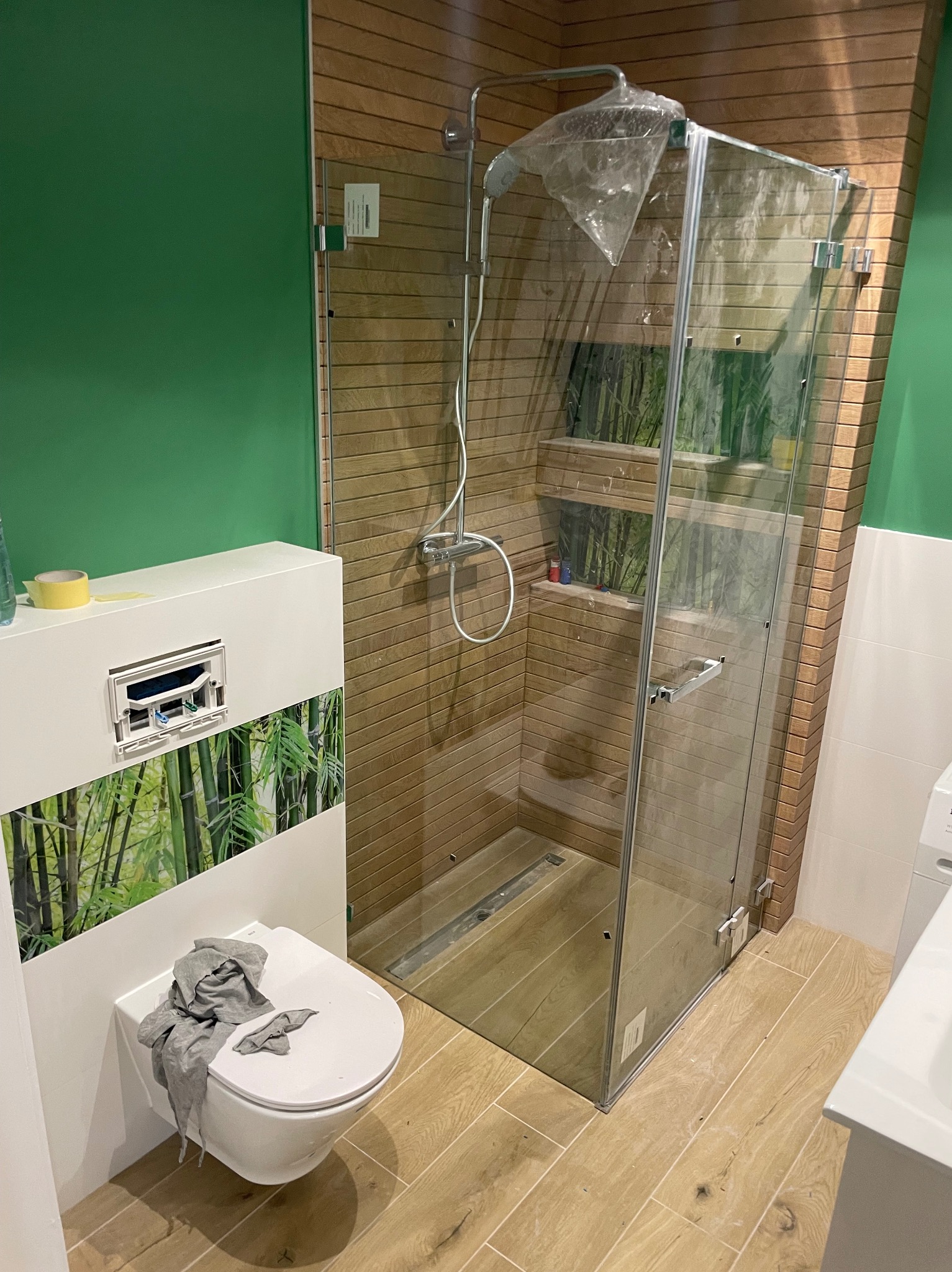 Nowoczesna łazienka w trakcie wykańczania: kabina prysznicowa z folią ochronną, podwieszana toaleta z motywem bambusa, drewnopodobne płytki na ścianach i podłodze, zielona ściana.