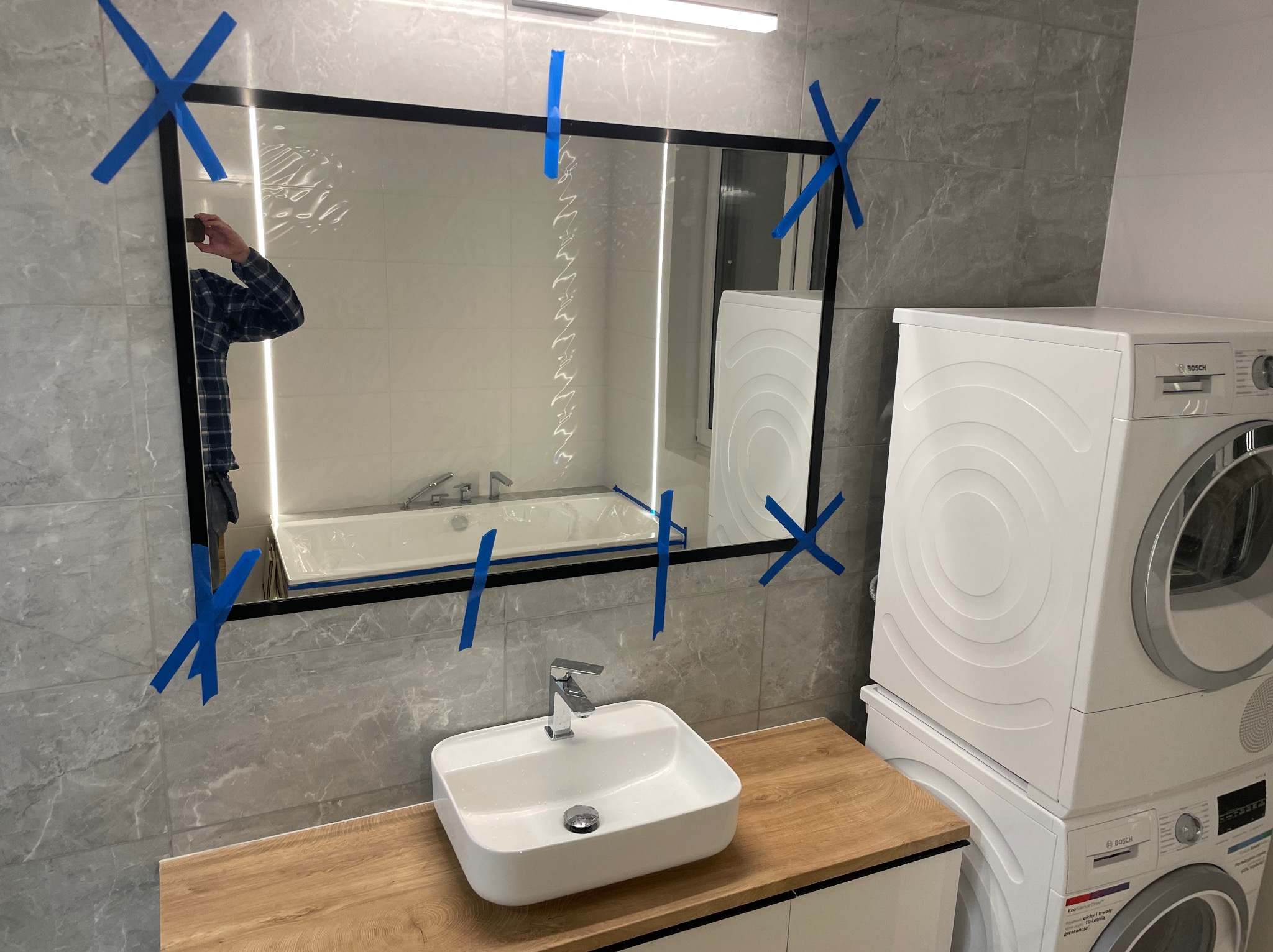 Nowoczesna łazienka z umywalką nablatową, lustrem w czarnej ramie zabezpieczonym taśmą malarską, pralką i suszarką Bosch w zabudowie; widoczne odbicie w lustrze przedstawia wannę i fragment osoby...