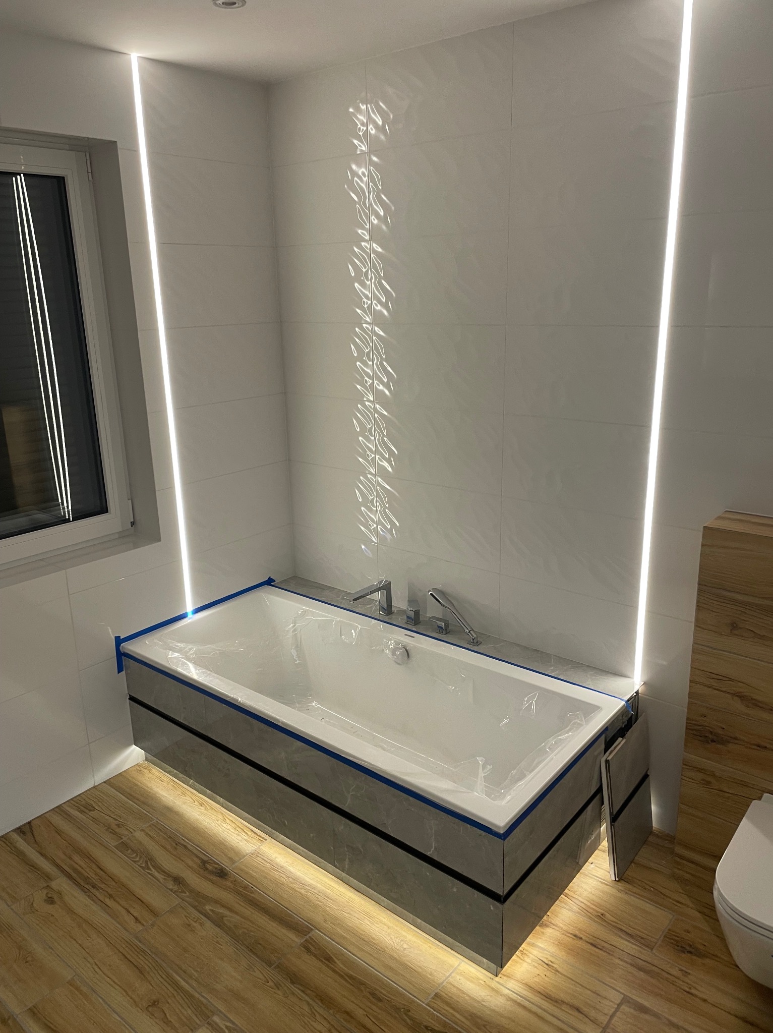 Wanna obudowana płytkami imitującymi marmur, zabezpieczona folią i niebieską taśmą malarską, oświetlona pionowymi listwami LED w nowoczesnej łazience z drewnopodobną podłogą i częściowo...