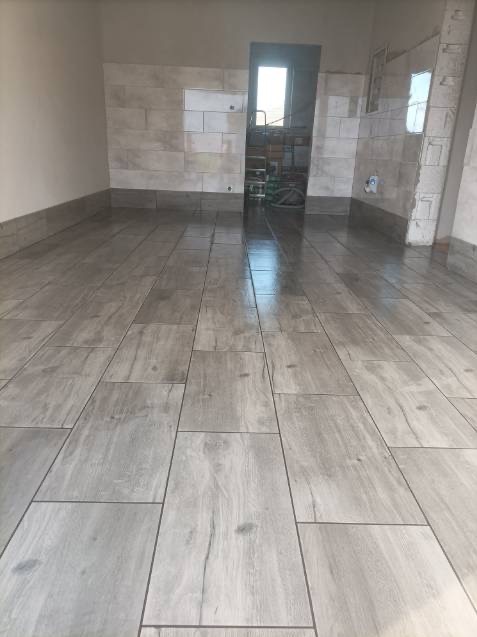 Ułożone podłużne płytki podłogowe imitujące drewno w jasnoszarym odcieniu, przy ścianach widoczne płytki ścienne, w głębi pomieszczenia otwarte przejście do innego pomieszczenia z widocznym sprzętem.