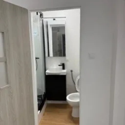 Nowoczesna łazienka po remoncie: kabina prysznicowa, umywalka z czarną szafką, toaleta i drewnopodobne płytki podłogowe. Widok zza uchylonych drzwi.