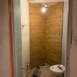 Wnętrze łazienki w trakcie remontu. Widoczne ułożone płytki imitujące drewno na ścianie z użyciem zielonych krzyżyków dystansowych, białe płytki, narzędzia glazurnicze.