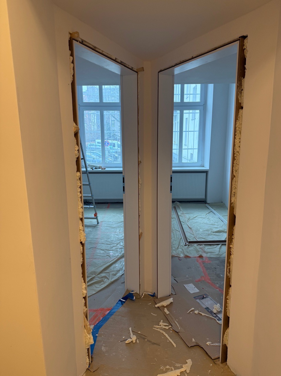 Przygotowanie do montażu ościeżnic w nowym otworze drzwiowym. Widoczne pianka montażowa, zabezpieczenia podłogi i drabina w tle. Prace remontowe w jasnym wnętrzu.