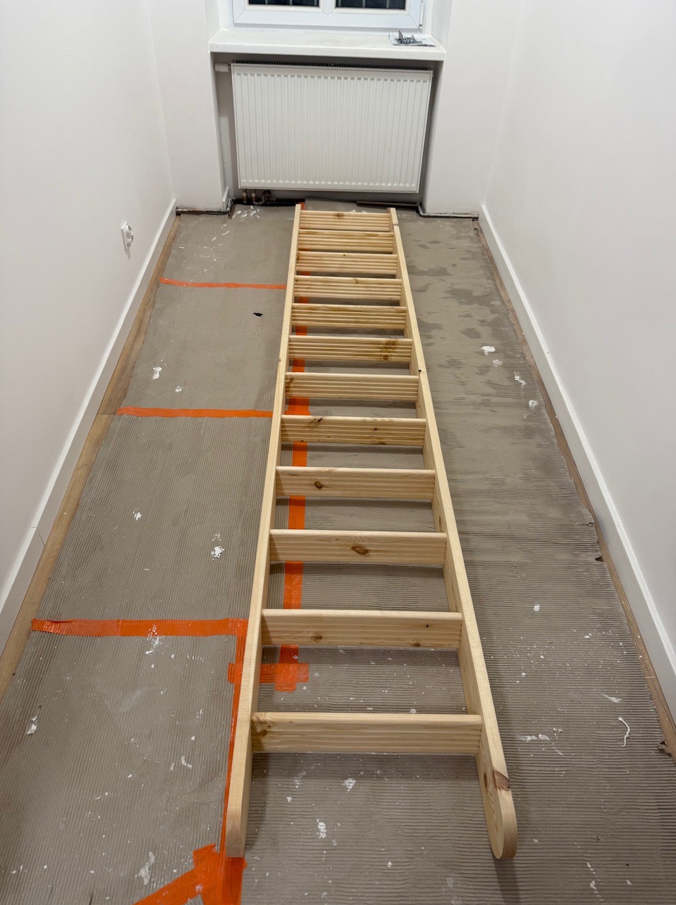 Przygotowanie podłoża pod płytki w wąskim korytarzu, z widocznym gruntem, taśmą malarską i drewnianą drabiną opartą o ścianę. Widok z góry.