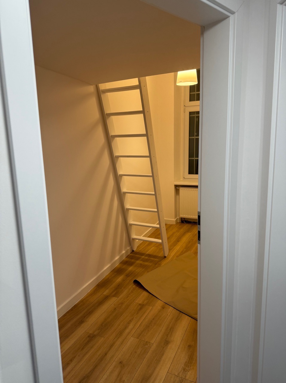 Biała drabina w pokoju z drewnianą podłogą, widoczna przez otwarte drzwi. Minimalistyczny design, ciepłe oświetlenie, fragment okna z grzejnikiem w tle.