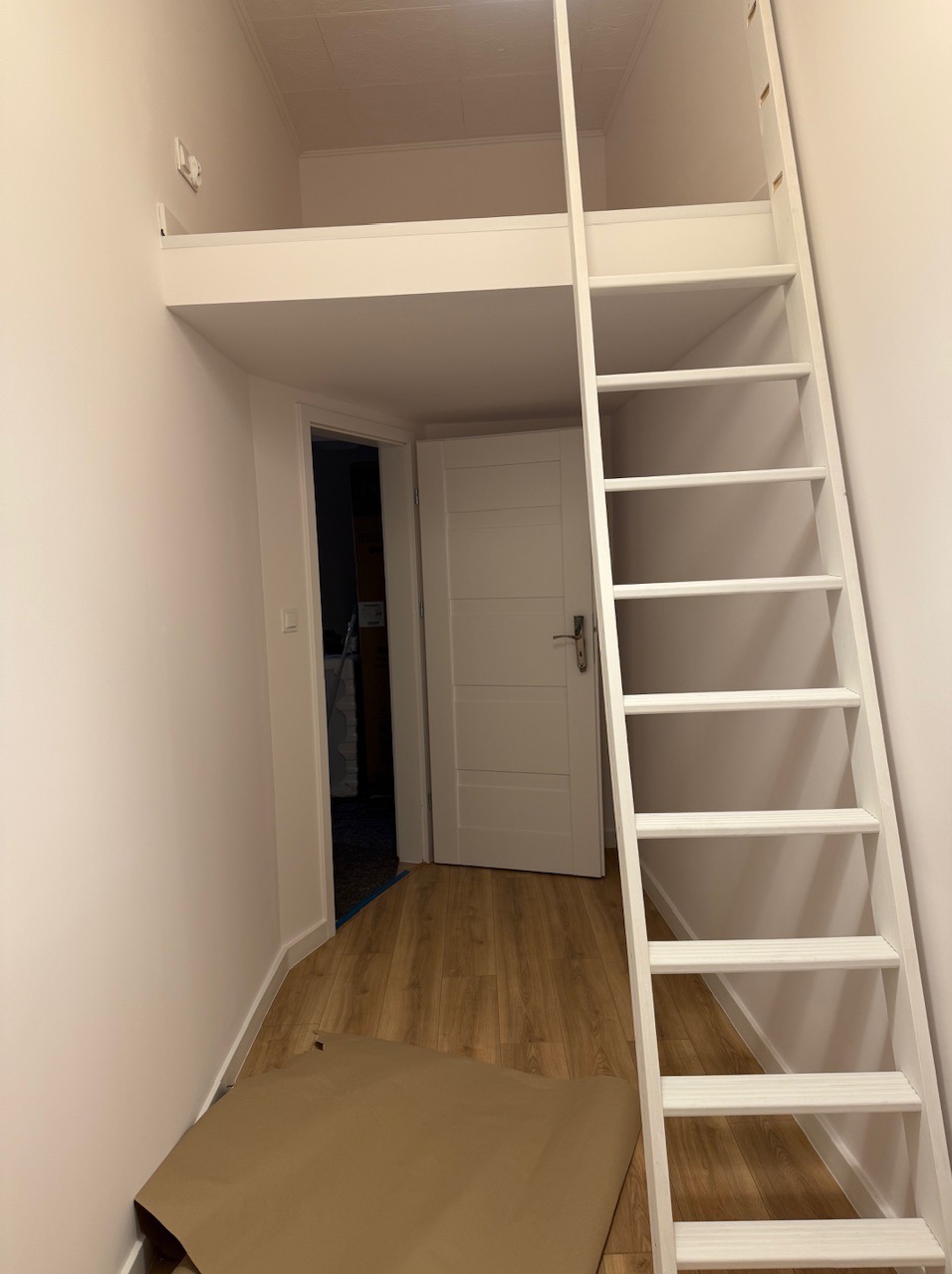 Biała drabina prowadząca na antresolę w pokoju z drewnianą podłogą. Widoczny fragment rozłożonego kartonu na podłodze. Otwarte drzwi do innego pomieszczenia.