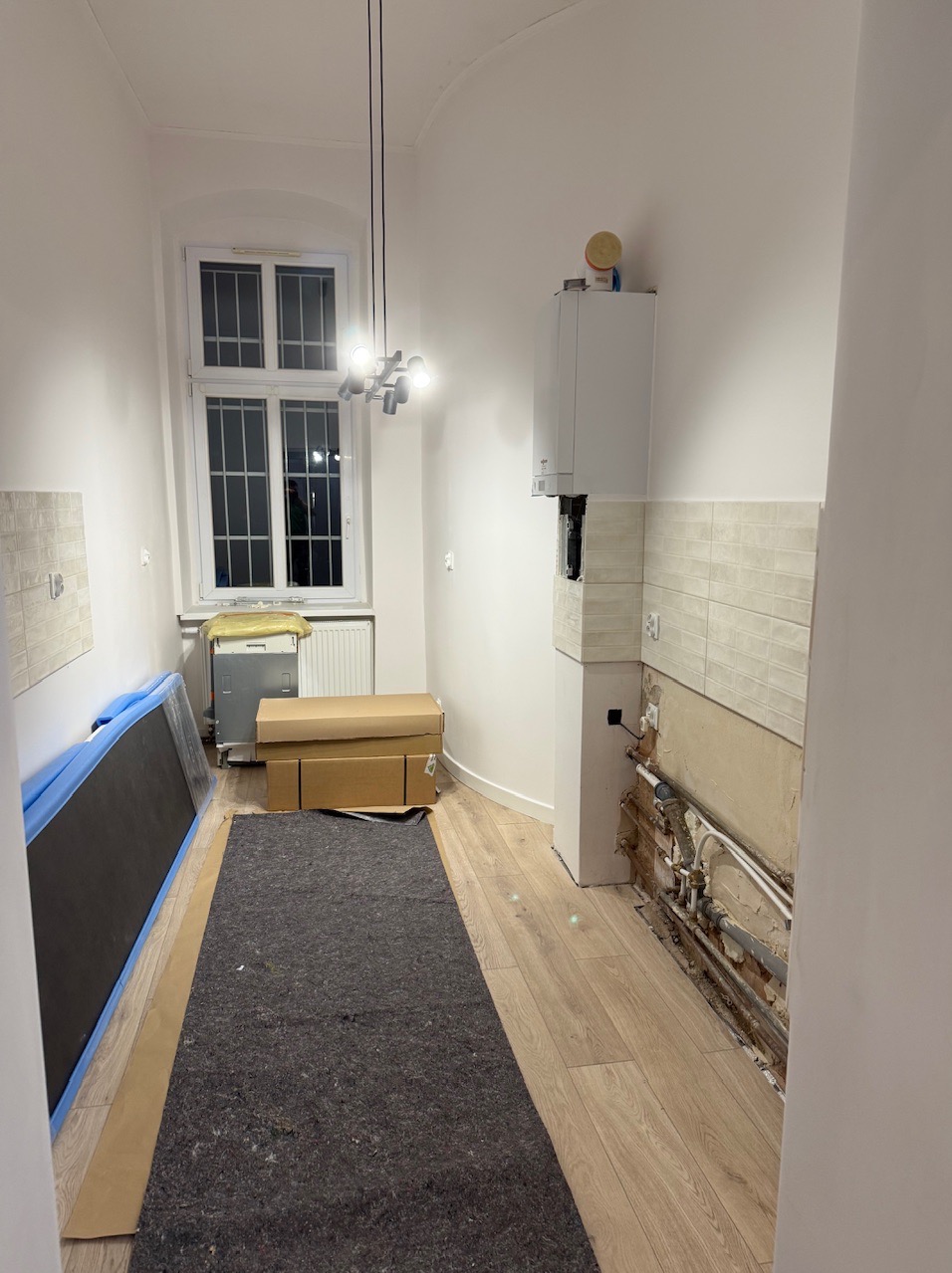 Wnętrze kuchni w trakcie demontażu płytek, z widocznymi rurami, zabezpieczonymi podłogami i przygotowanym miejscem pod zabudowę. Nowoczesne oświetlenie.