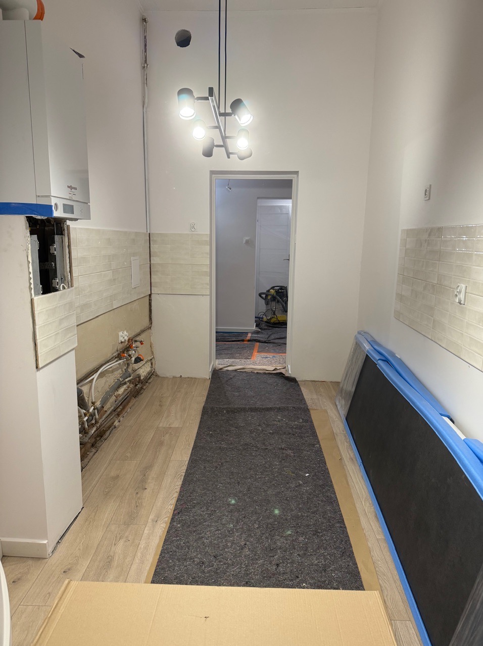 Wnętrze kuchni w trakcie remontu. Widoczne fragmenty ułożonych płytek, odsłonięte rury, zabezpieczona podłoga oraz sprzęt. Częściowo wykończone ściany i oświetlenie.