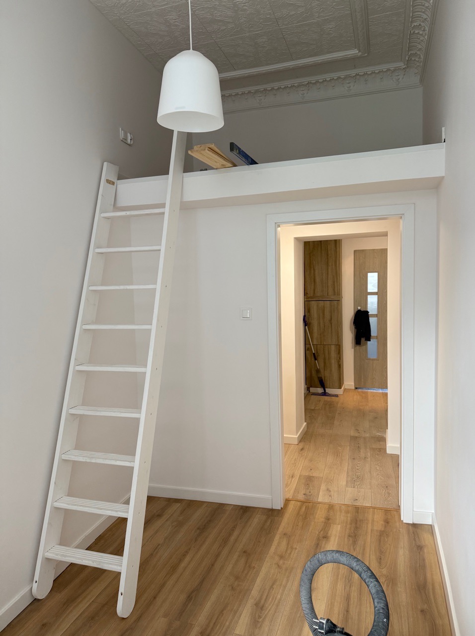 Białe schody prowadzące do antresoli w pokoju z parkietem. Widoczna lampa i fragment sufitu. W tle drzwi do innego pomieszczenia.