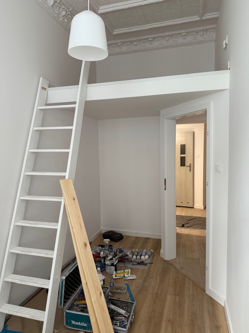 Pokój po remoncie z antresolą i białą drabinką. Widoczne narzędzia i materiały malarskie. W tle jasne podłogi i drzwi do innego pomieszczenia.