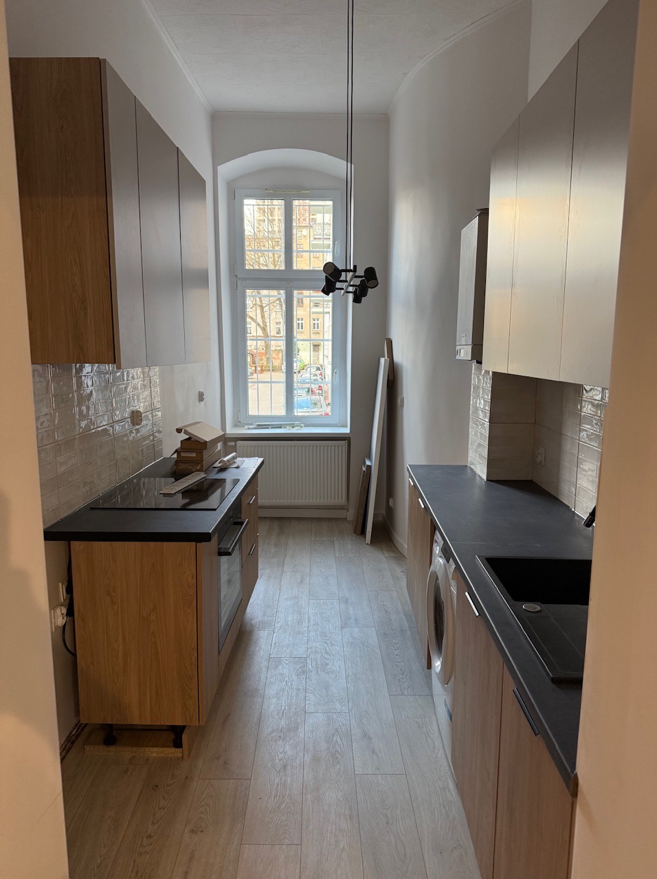 Wąska kuchnia po remoncie z drewnianymi szafkami, czarnym blatem i jasną podłogą. Okno z widokiem na ulicę doświetla wnętrze. Nowoczesna lampa sufitowa.