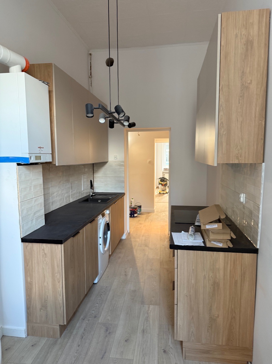 Nowoczesna kuchnia w trakcie wykończenia: jasne szafki, czarny blat, płytki i sprzęt AGD. Widoczne kartony i plany.