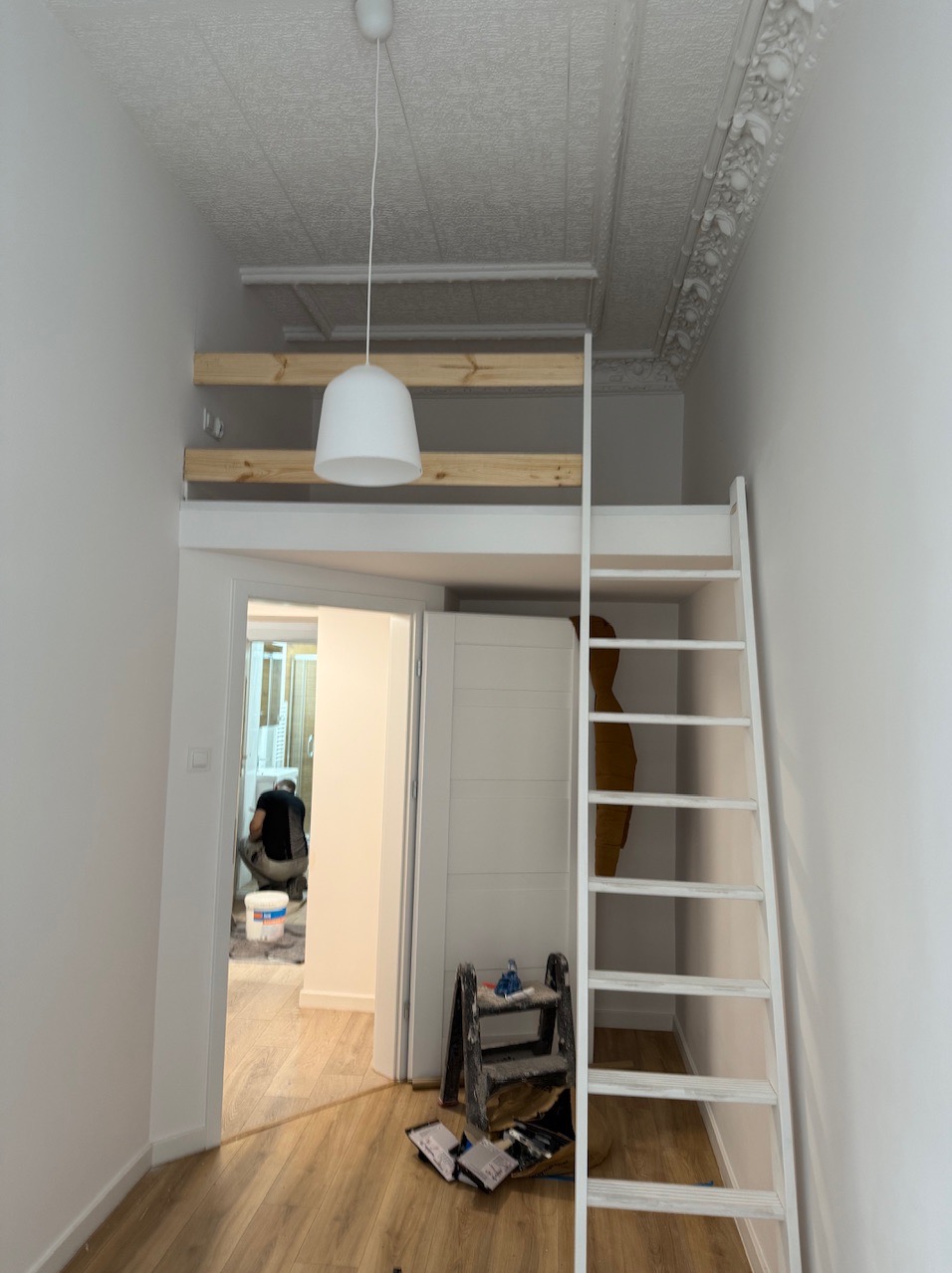 Wnętrze po remoncie: białe ściany, drewniana podłoga, antresola z drabinką. W tle malarz przy pracy, puszki z farbą. Dekoracyjny sufit z gzymsem.