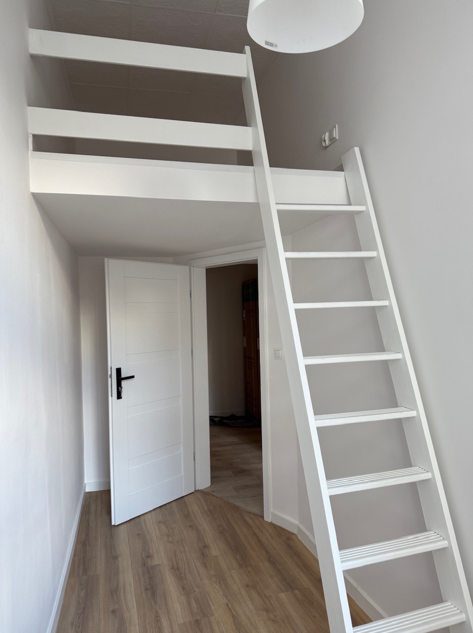 Białe łóżko na antresoli z drabinką w pokoju. Widoczne drewniane panele podłogowe i otwarta biała  drzwi. Minimalistyczny design i jasne wnętrze.