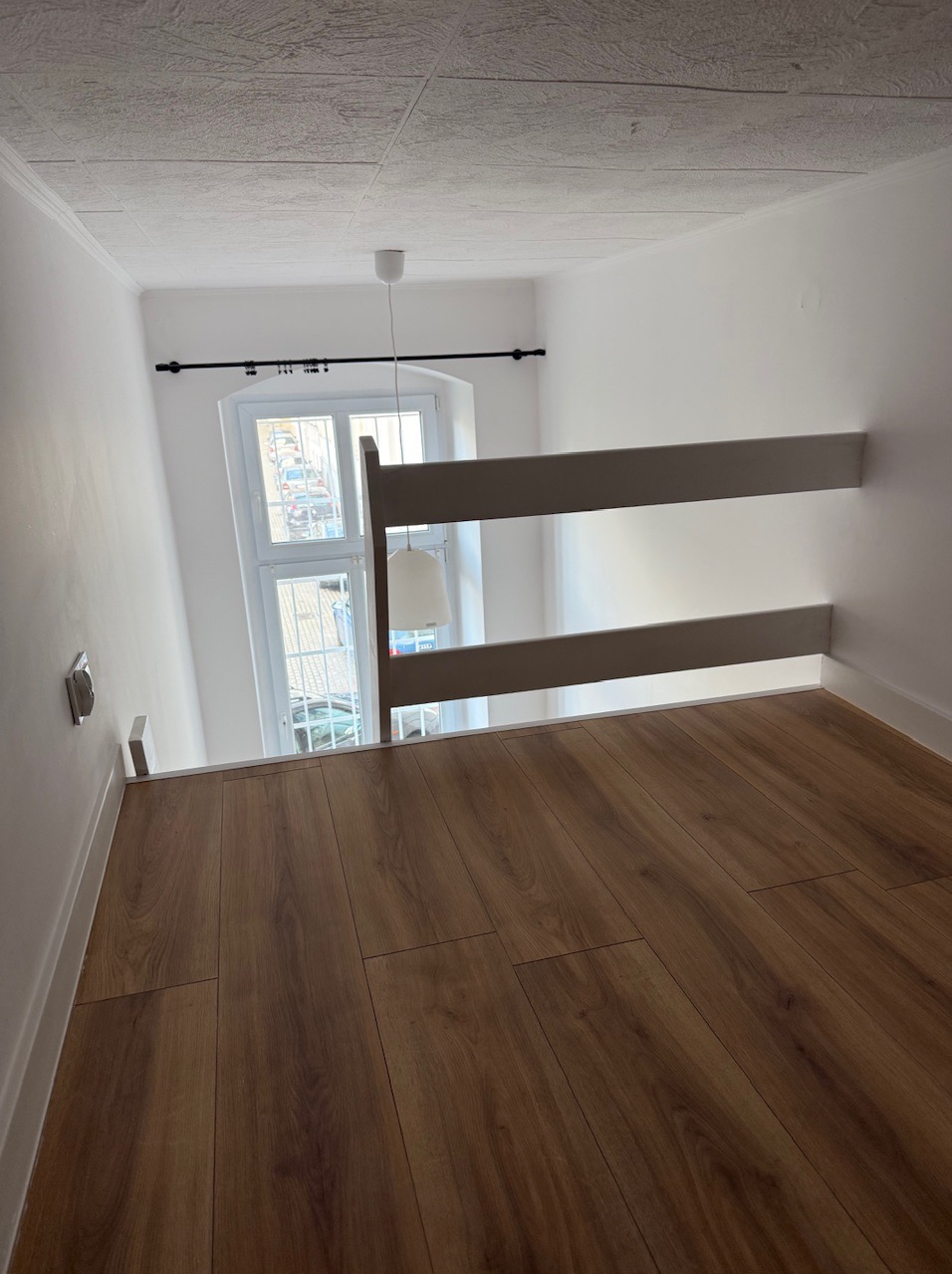 Jasne panele podłogowe w pokoju z antresolą i oknem. Widok z góry, delikatne oświetlenie. Minimalistyczne wnętrze.