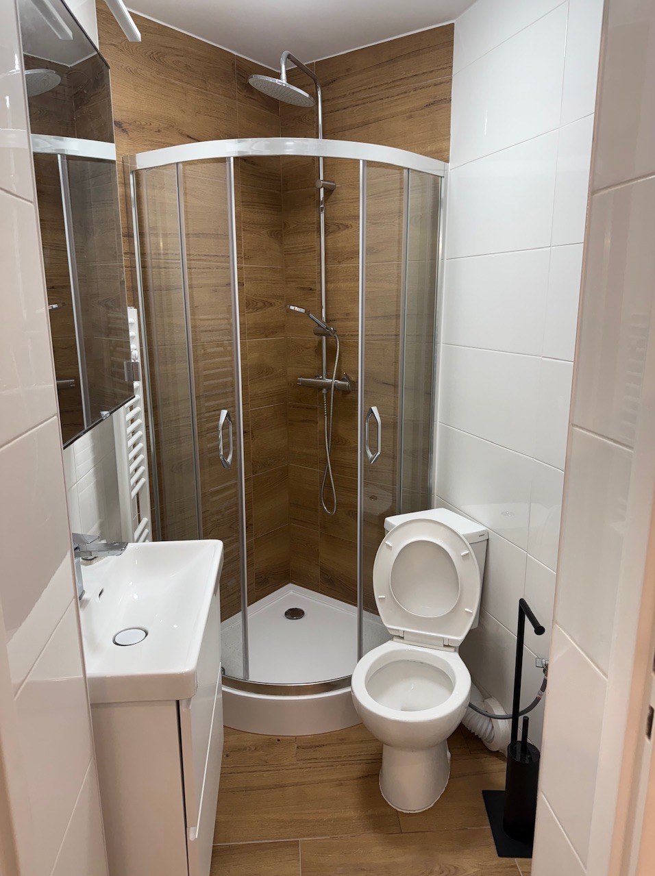 Nowoczesna łazienka po remoncie: kabina prysznicowa narożna z drewnopodobnymi płytkami, biała umywalka z szafką i toaleta. Wykończenie w bieli i drewnie.