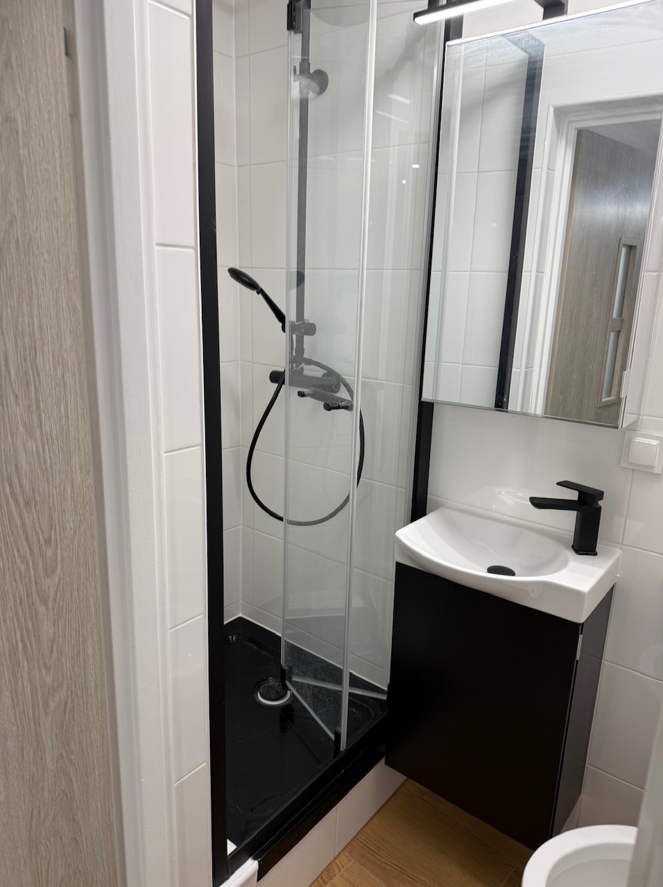 Nowoczesna kabina prysznicowa z czarnymi profilami i armaturą. Umywalka z szafką w kolorze czarnym, białe płytki na ścianach, jasna podłoga. Minimalistyczny design.