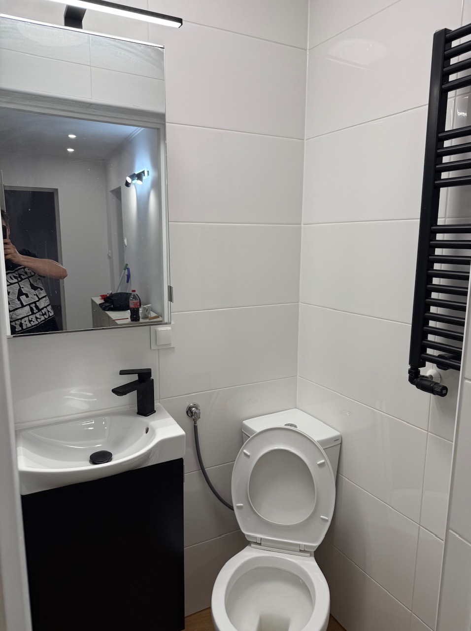 Nowoczesna, biała łazienka po remoncie. Umywalka z czarną armaturą, toaleta z otwartą klapą, grzejnik drabinkowy. Widoczne białe płytki na ścianach.