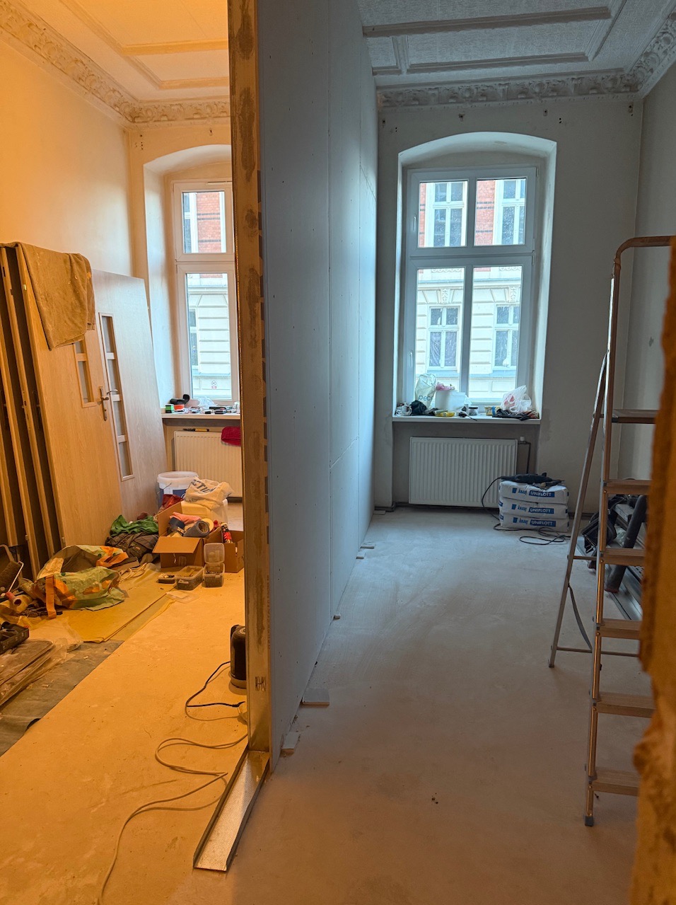 Remont mieszkania w kamienicy: widok na pokój z oknem i grzejnikiem, ściana z płyty gipsowej w trakcie montażu, drzwi i drabina w tle.