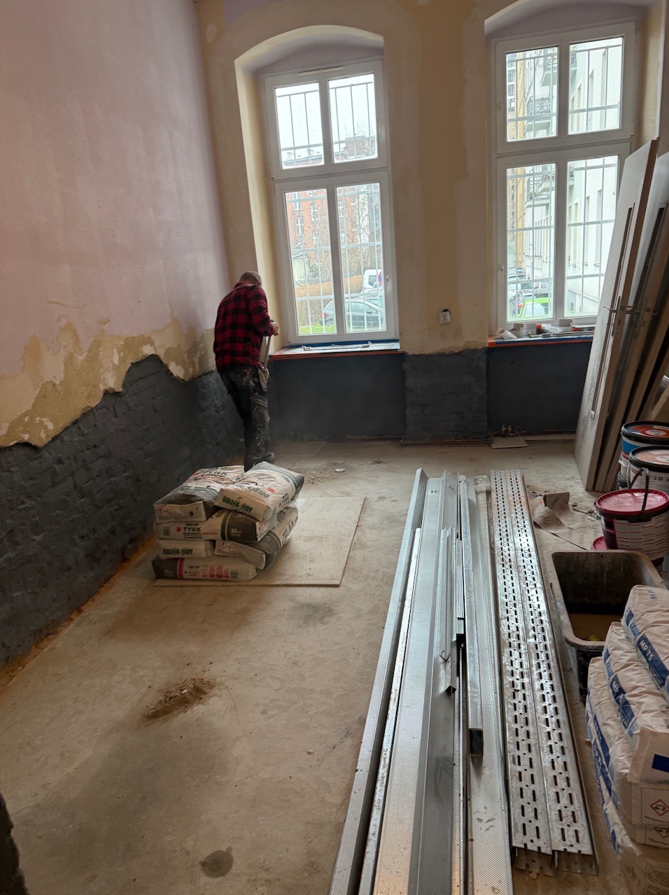 Remont pokoju: murarz w koszuli w kratę obrabia ścianę przy oknie, obok worki z zaprawą i profile. Etap przygotowania ścian do dalszych prac wykończeniowych.