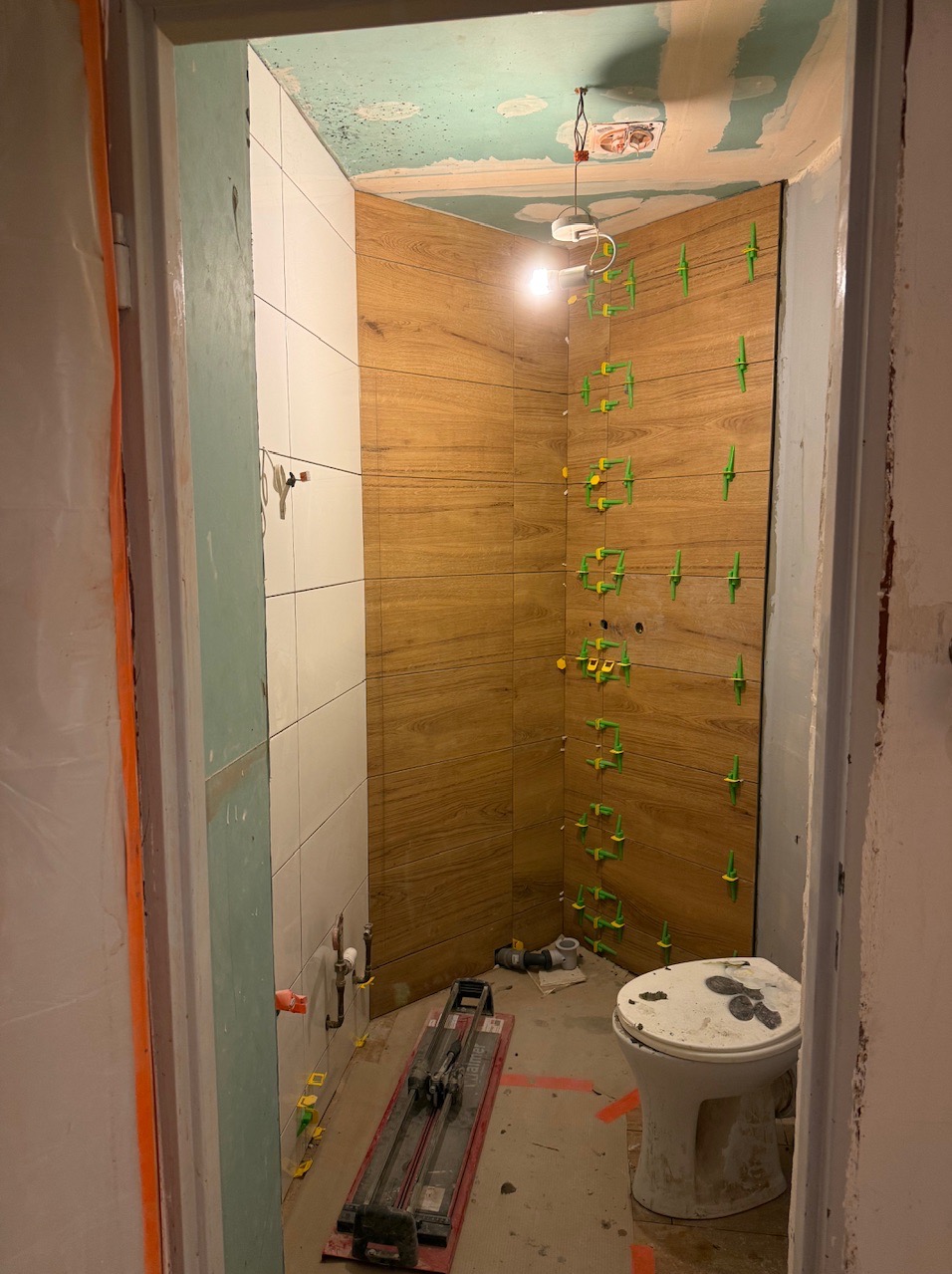 Wnętrze łazienki w trakcie remontu. Widoczne ułożone płytki imitujące drewno na ścianie z użyciem zielonych krzyżyków dystansowych, białe płytki, narzędzia glazurnicze.