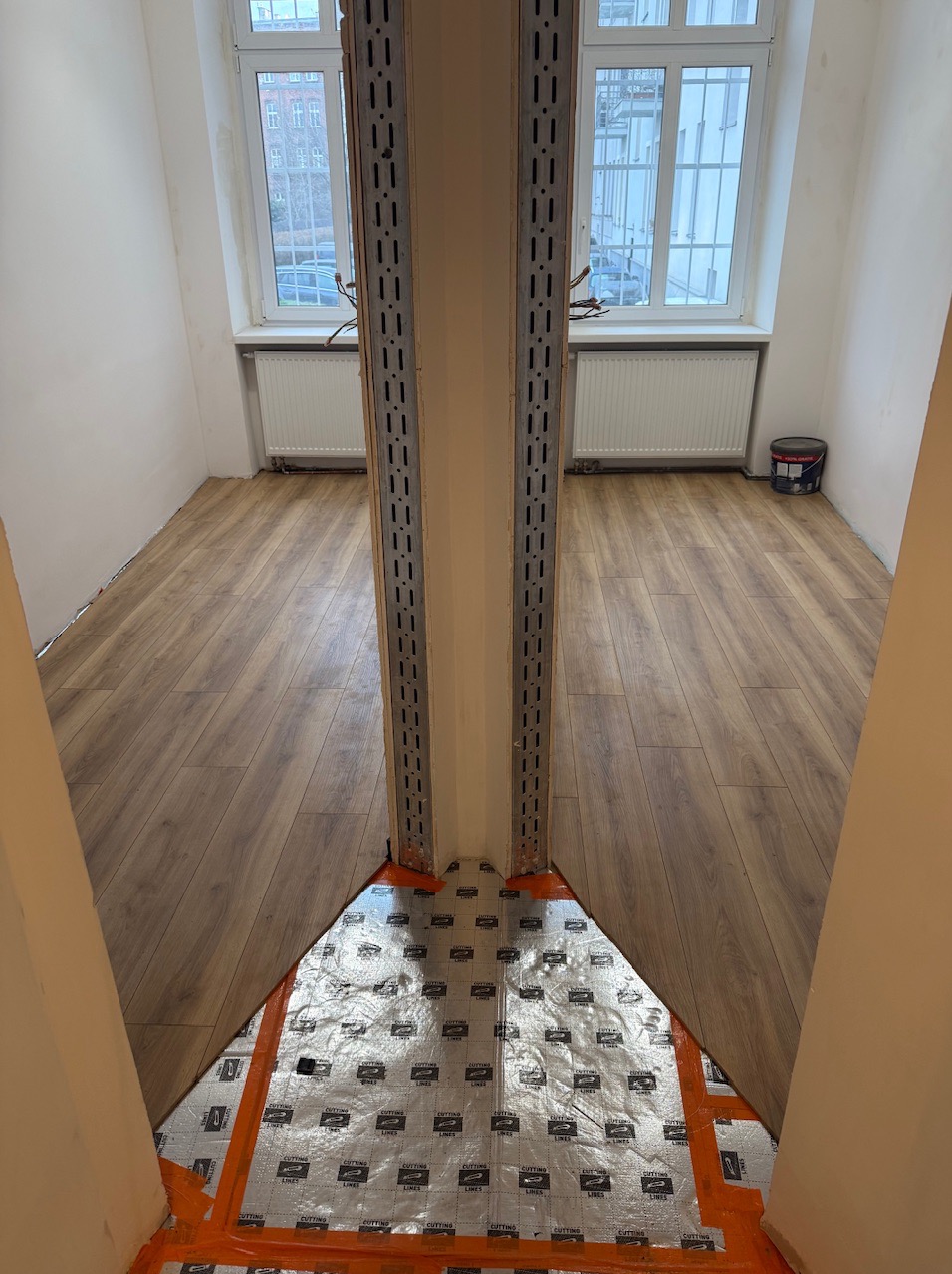 Montaż profili i izolacji pod panele podłogowe w jasnym pokoju z oknem. Widoczne elementy instalacji i wykończona podłoga.