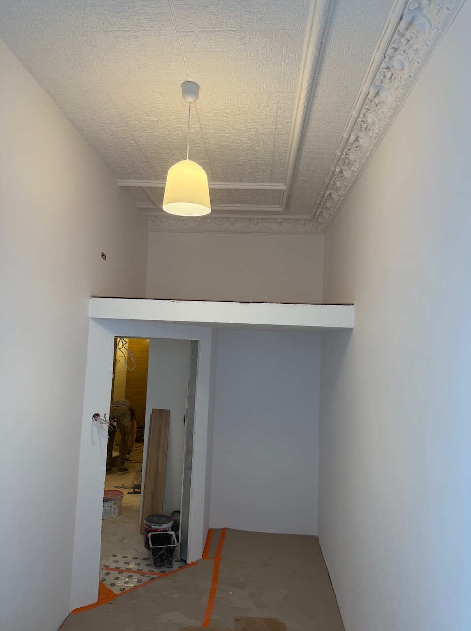 Wnętrze w trakcie remontu: białe ściany, ozdobny sufit z żyrandolem, widoczne przygotowania do prac wykończeniowych i narzędzia w tle.
