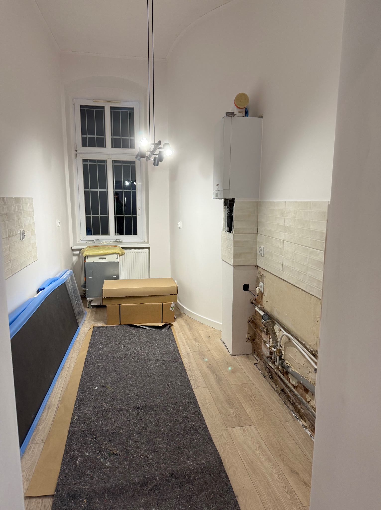 Wnętrze w trakcie remontu: odsłonięte rury, zabezpieczona podłoga panelami, kartony i sprzęt pod oknem. Częściowo położone płytki ścienne.