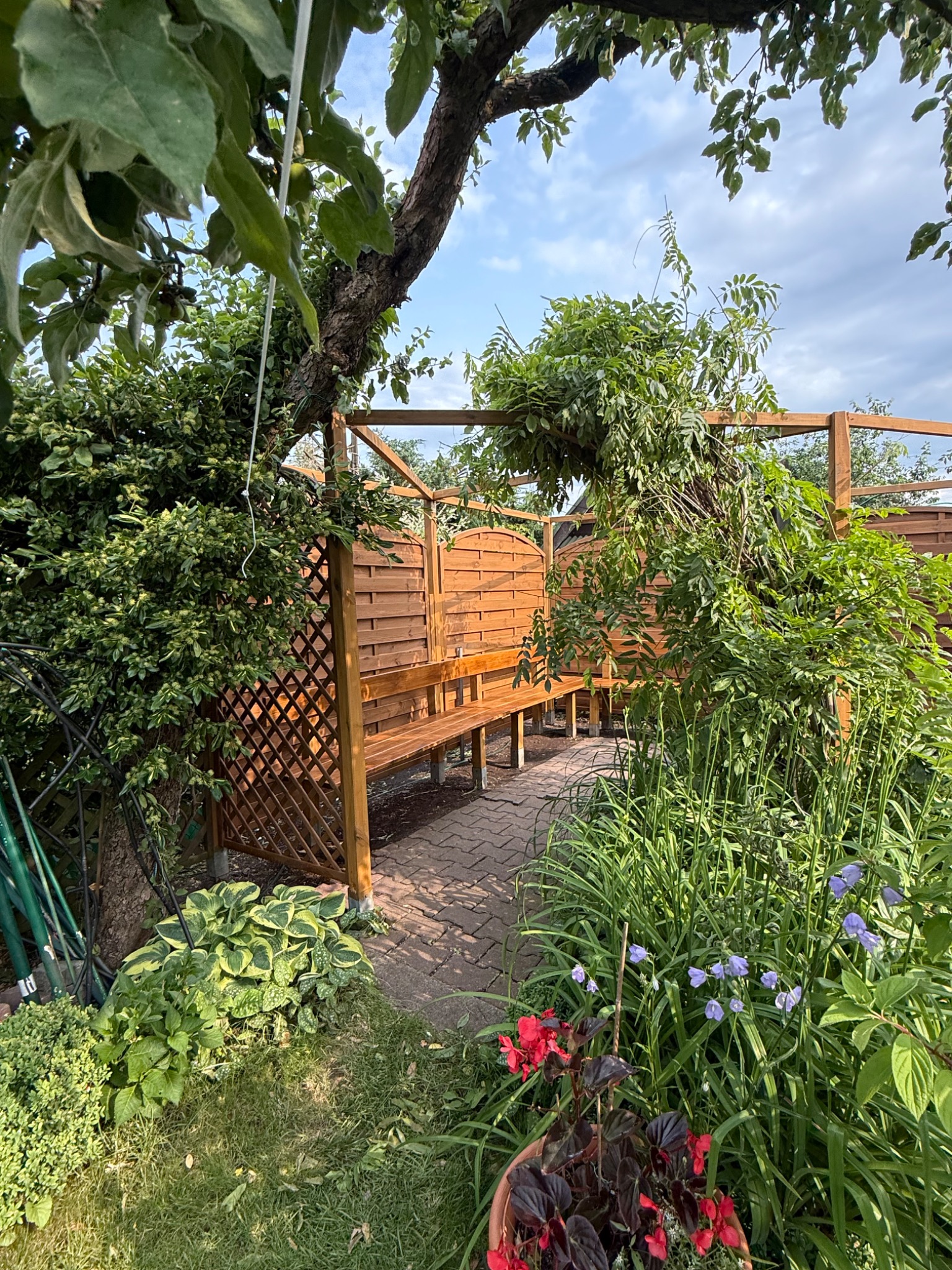 Drewniana altana z ławką, porośnięta zielenią, tworząca zaciszne miejsce w ogrodzie. Widoczne kwiaty i ścieżka z kostki brukowej. Całość w naturalnej, letniej scenerii.