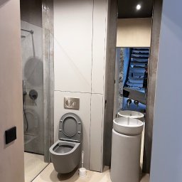 KERAM-BUD - Nowoczesna łazienka w odcieniach szarości z toaletą, umywalką na postumencie i przeszklonym prysznicem; w lustrze odbicie schodów zabezpieczonych folią malarską.