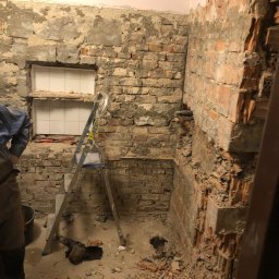 KERAM-BUD - Demontaż starej ściany w łazience, odsłonięta cegła, gruz na podłodze, drabina aluminiowa, widoczne rury i fragment okna z białymi płytkami w tle.