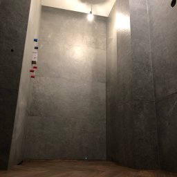 KERAM-BUD - Wnętrze łazienki w trakcie prac wykończeniowych; szare płytki imitujące kamień na ścianach, drewniana podłoga ułożona w jodełkę, widoczne wyprowadzenia instalacji wodnej i elektrycznej, surowa...