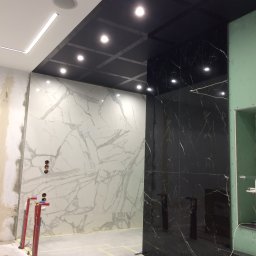 KERAM-BUD - Wnętrze w trakcie prac wykończeniowych: ściany z białych i czarnych płyt imitujących marmur, widoczne instalacje wodne i fragment sufitu z oświetleniem punktowym.