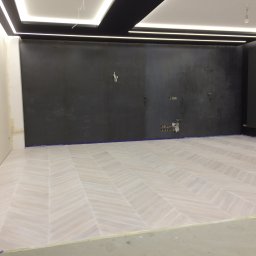 KERAM-BUD - Jasna podłoga ułożona w jodełkę w pokoju z czarną ścianą i nowoczesnym oświetleniem sufitowym, widoczne przygotowania do dalszych prac wykończeniowych.