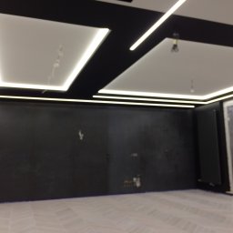KERAM-BUD - Wnętrze pokoju w trakcie prac wykończeniowych, z czarną ścianą, białymi panelami sufitowymi z oświetleniem LED i podłogą z jasnych płytek imitujących drewno; widoczne niedokończone instalacje...