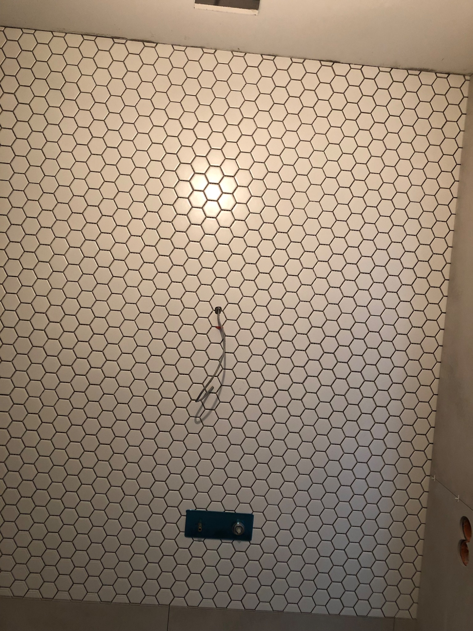 Ściana łazienki wyłożona białymi, heksagonalnymi płytkami z ciemną fugą, widoczny wystający przewód i zamontowany przycisk spłuczki w kolorze turkusowym.