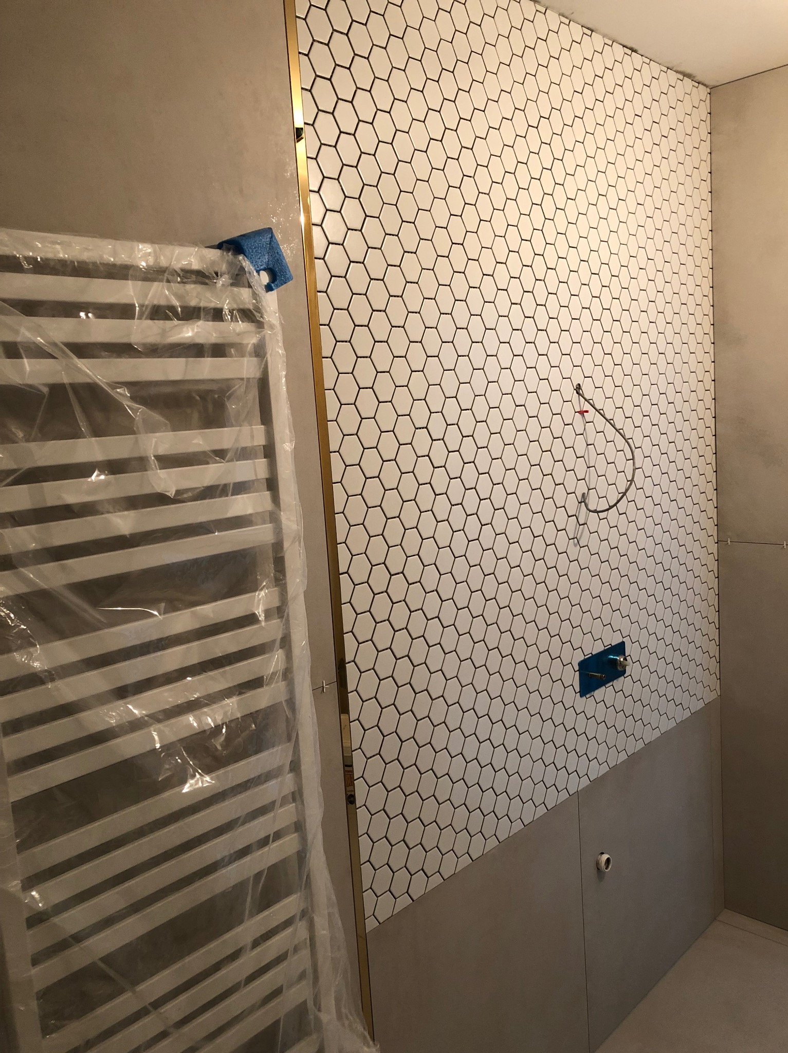 Ściana łazienki wyłożona białymi heksagonalnymi płytkami z czarną fugą, zakończona złotą listwą. Na ścianie widoczne przygotowane przyłącza wodne. Grzejnik drabinkowy owinięty folią ochronną po...