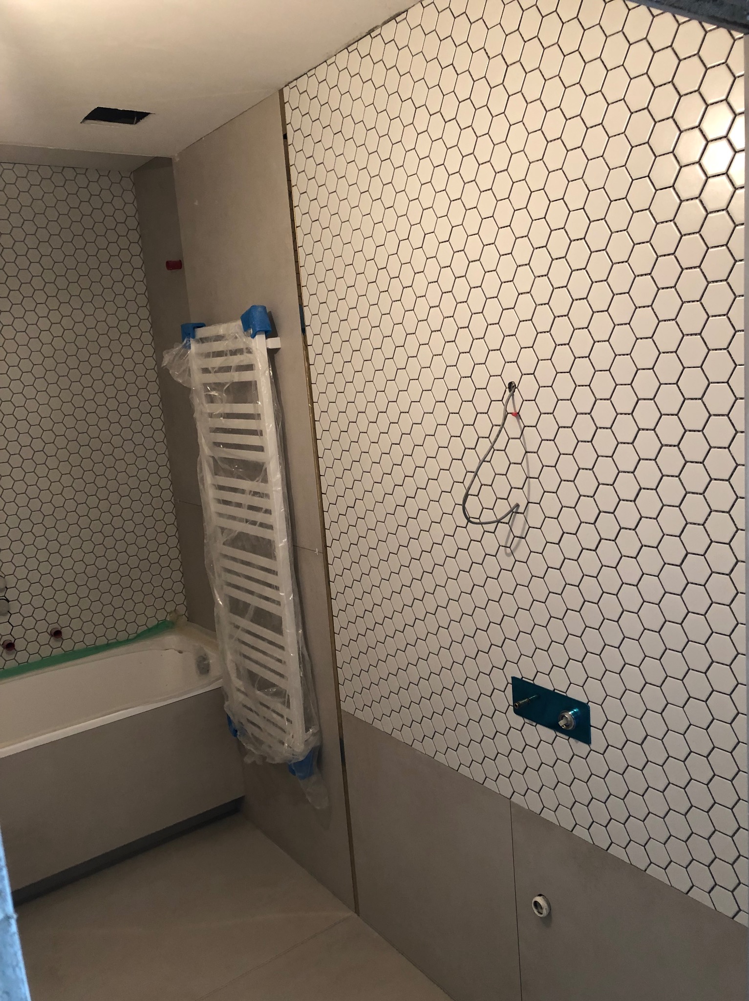 Wnętrze łazienki w Krakowie w trakcie prac wykończeniowych: białe heksagonalne płytki na ścianie, grzejnik drabinkowy w folii ochronnej, widoczna instalacja wodna i fragment wanny.