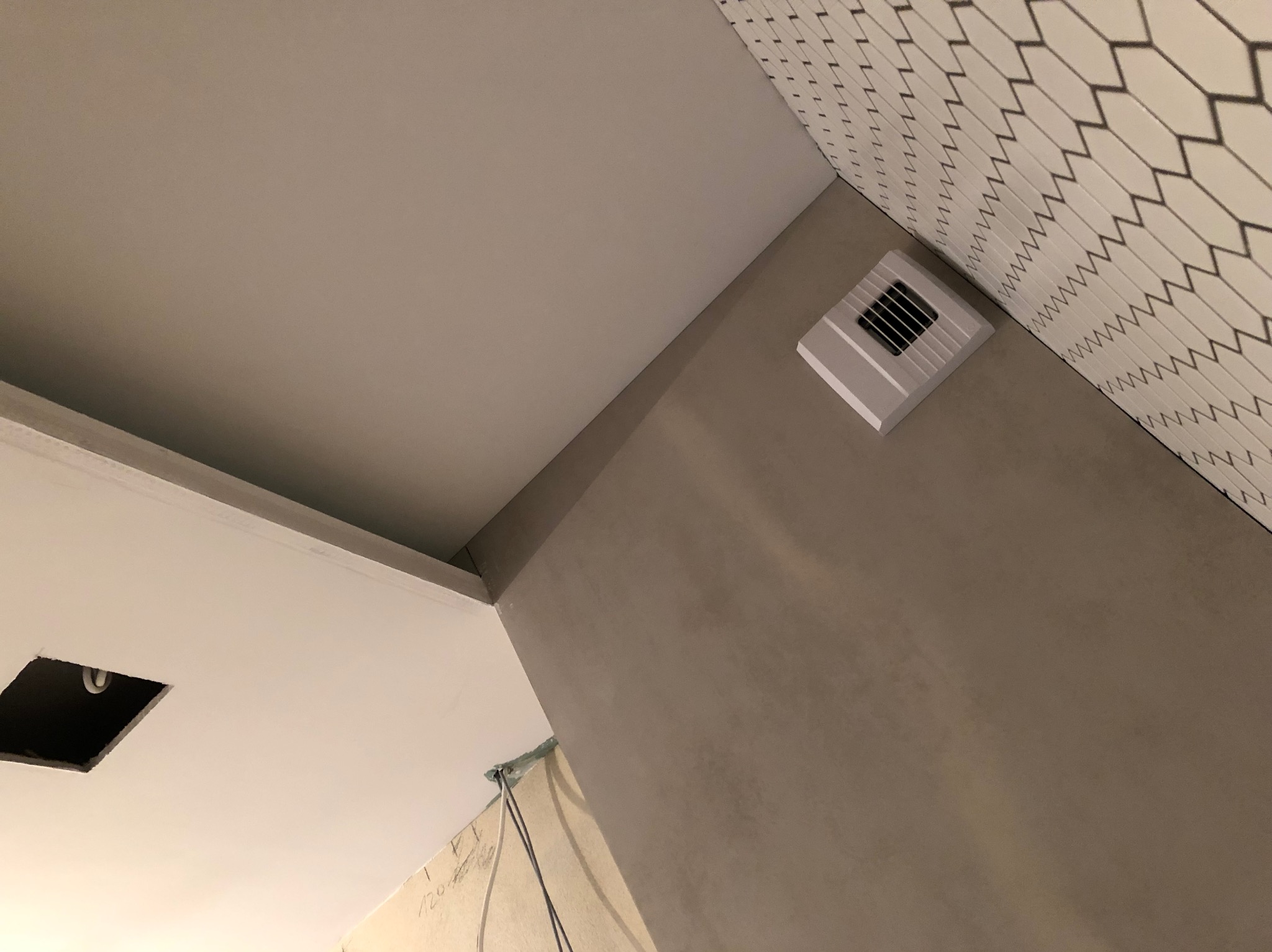 Narożnik łazienki w trakcie remontu: ściana z szarą fakturą, fragment sufitu z otworem na oświetlenie, wentylator na ścianie i fragment ściany wyłożonej białymi płytkami z czarnym wzorem w romby.